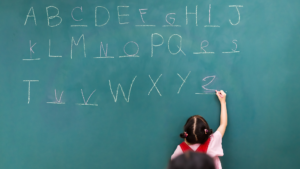 The letters of the alphabet are written out on a chalkboard, but there are blanks in place of a few of the letters. A school girl is writing in the missing letters.